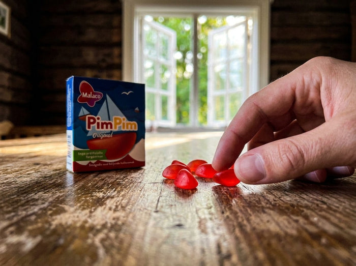 Malaco PimPim Original Tablettask 20g open blue and red box beside five small red boat-shaped raspberry gummy candies on a rustic wooden table, hand picking one up, summer window in background