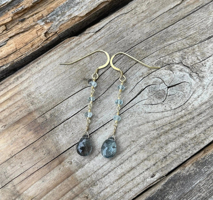 Alice Gemstone Earrings | Liv & B featuring delicate gold hooks with light blue gemstone beads and teardrop-shaped blue gemstones hanging on a rustic wooden background.