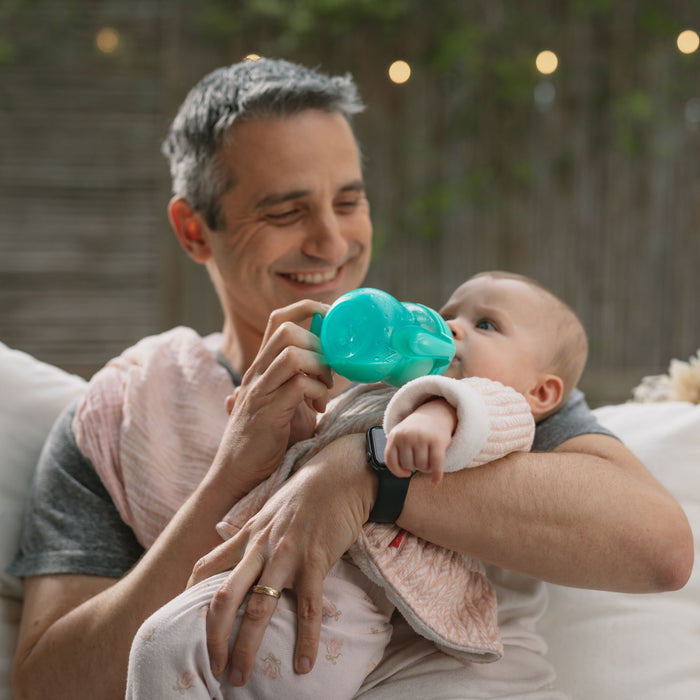 A smiling man in a wedding ring and smartwatch feeds a baby dressed in pink with the Bottle-to-Cup Wide Neck Bottle, featuring a green no-spill spout, as they sit together on a couch against a softly blurred background.