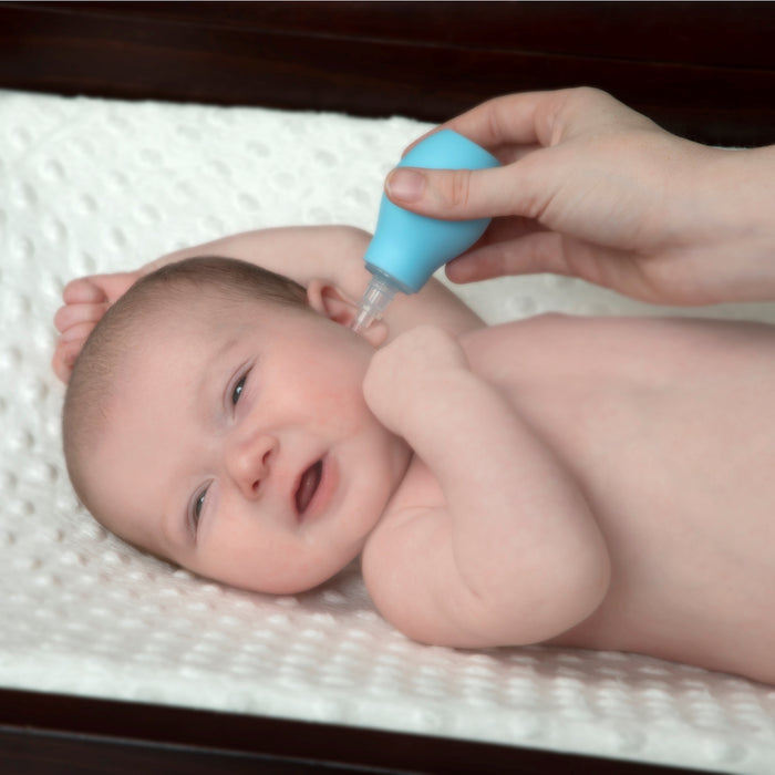 A baby with one arm raised lies on a soft, white surface while an adult uses the blue bulb from the Nuby Medical Kit to clean the baby's ear. The baby's eyes are partially open.