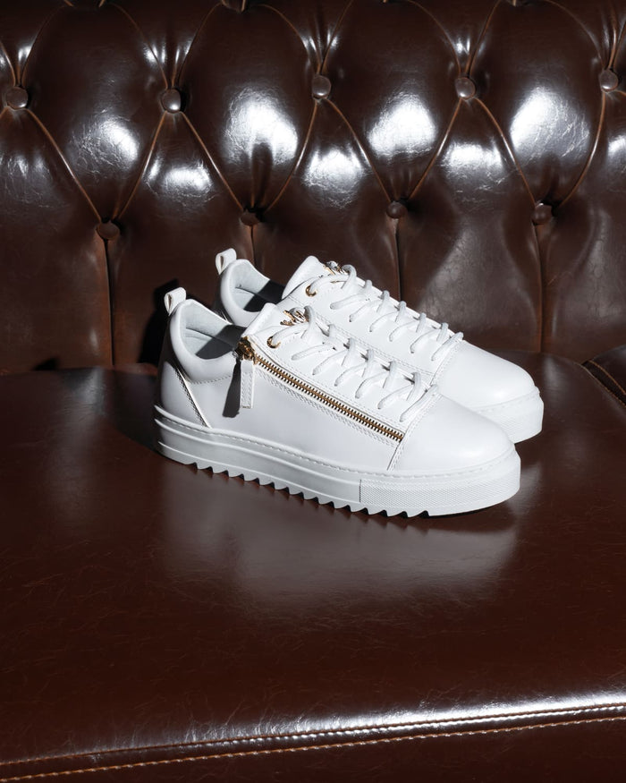 Martin Valen men's low-top white sneakers with gold zipper, front three-quarter view displayed on dark leather seat