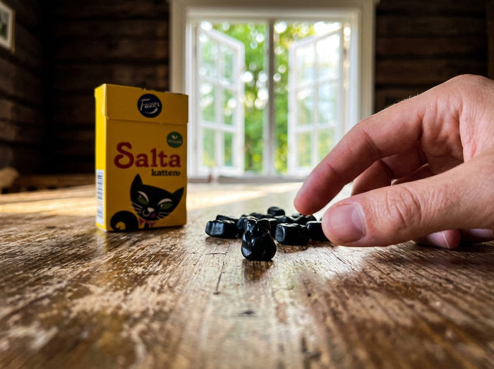 Fazer Salta Katten Tablettask 38g open yellow box beside a pile of small black cat-shaped salmiak licorice pastilles on a rustic wooden table, hand picking up one pastille, summer window in background