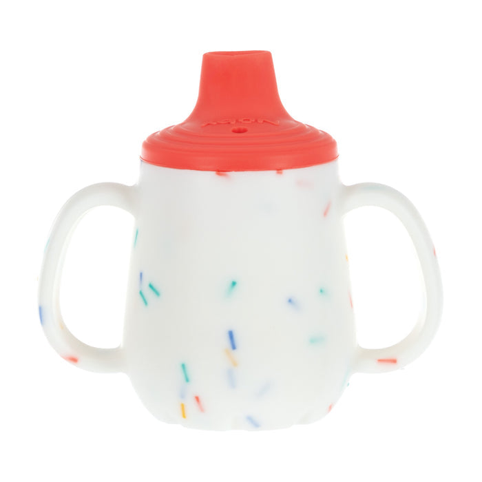 The Nuby US 2-Handle First Training Cup | Red Confetti is a white silicone baby cup with two handles, a red lid, and confetti prints—ideal as a first training cup with a free-flow spout for easy sipping.