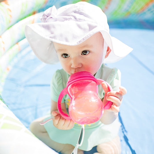 A baby in a wide-brimmed white hat sits in a small inflatable pool, enjoying sunshine while drinking from the 3-Stage Grow With Me Wide Neck Bottle, which features an anti-colic air system for a smooth transition from bottle to cup.