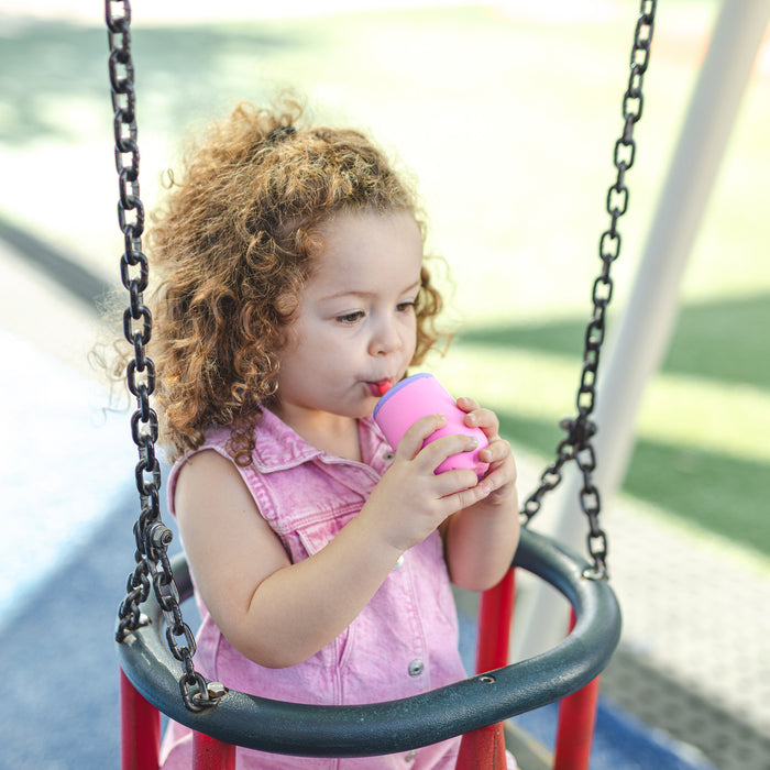 A young child with curly hair enjoys a sunny playground, wearing a pink sleeveless shirt and sipping from the 3-Stage Training Cup Set | Aqua, a no-spill straw cup. The background features green flooring in an outdoor play area.