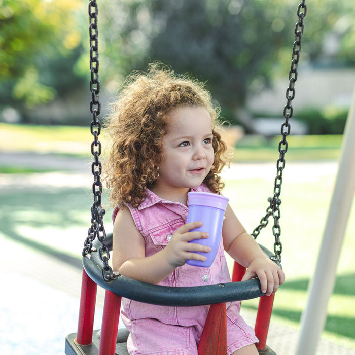 A young child in a pink dress sits on a playground swing, holding the 360 Wonder Cup (3 Pack - 10 oz, Purple/Pink/Aqua). Greenery and blurred outdoor elements fill the background.