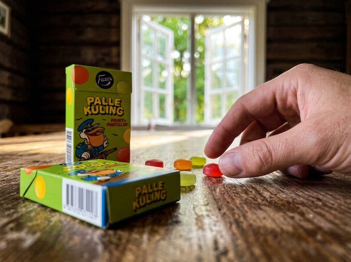 Fazer Palle Kuling Tablettask 38g open green box on a rustic wooden table with colorful soft fruit pastilles scattered beside it, hand picking up a red raspberry pastille, summer window with green trees in background