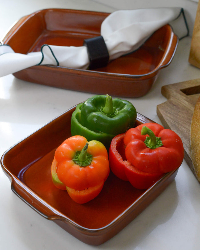 Three bell peppers (red, green, and orange) on a caramel ceramic stoneware baking dish with a white linen in the background.