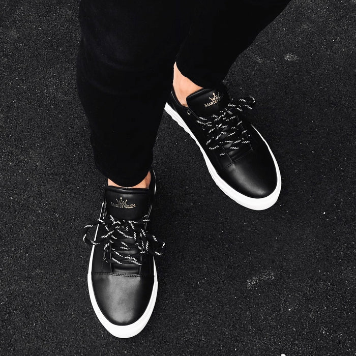 Martin Valen Artestitch black low-top sneakers worn, top-down view on asphalt with gold logo on the tongue