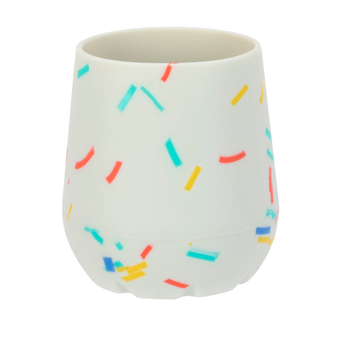 The Nuby US Baby's First Mini Sipper Training Cup is a white silicone cup with a matte finish, decorated with blue, yellow, orange, and red confetti sprinkles—ideal for open cup drinking training.