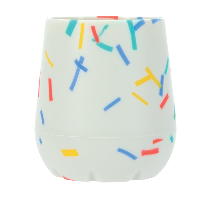 The Nuby US Baby's First Mini Sipper Training Cup is a white silicone cup with a curved design and matte finish, decorated with blue, red, yellow, and green confetti sprinkles—perfect for open cup training.