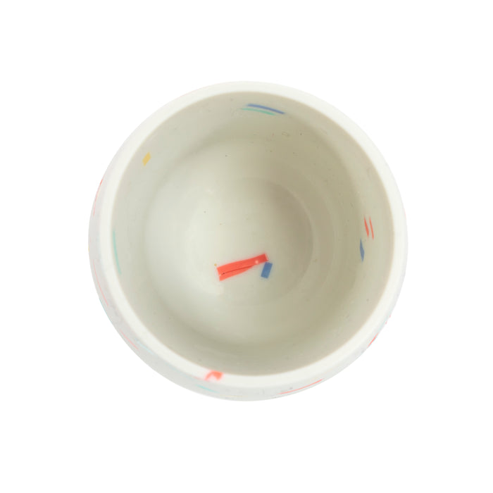 Top-down view of Nuby US Baby's First Mini Sipper Training Cup—a small white silicone cup with red, blue, yellow, and teal confetti brushstroke details, ideal for introducing open cup drinking.