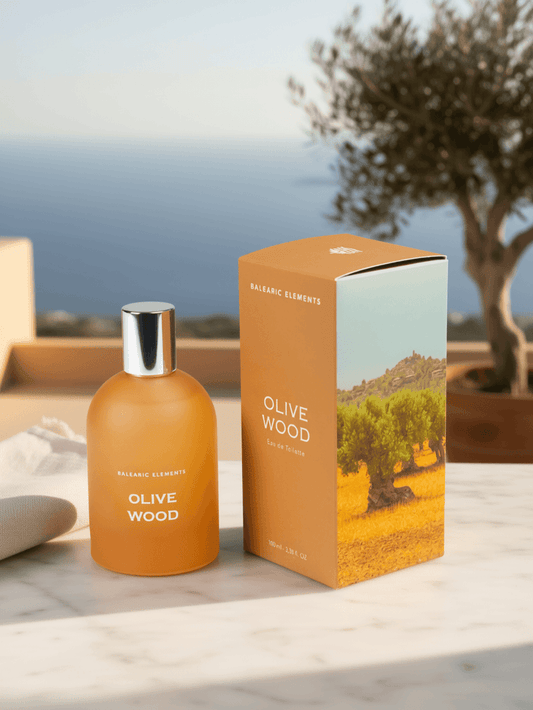 An amber glass bottle and matching orange box of Balearic Elements Olive Wood fragrance, displayed on a marble surface with a blurred outdoor background of trees and water.