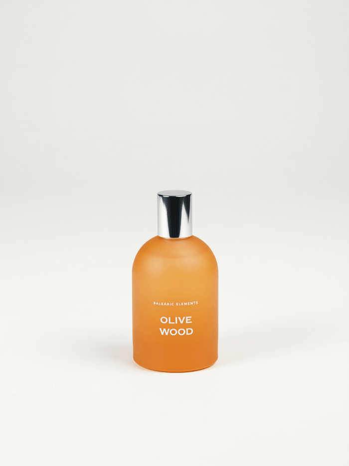 An orange frosted glass bottle with a shiny silver cap, labeled 'OLIVE WOOD' by Balearic Elements, set against a plain white background.