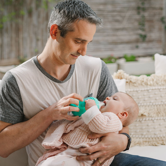 A man with short gray hair sits on a couch, gently feeding a baby in pink with the Bottle-to-Cup Wide Neck Bottle, featuring a no-spill spout. Neutral cushions and greenery decorate the background as the baby gazes up at him.