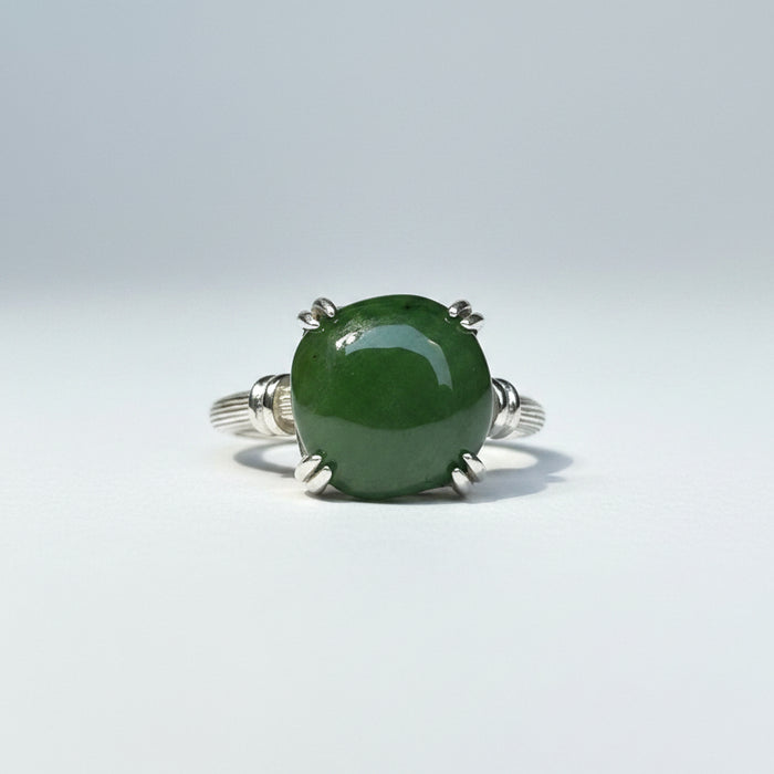 A silver ring with a large, oval green stone set in a four-prong setting, resting on a textured wooden surface with a soft-focus gray background.