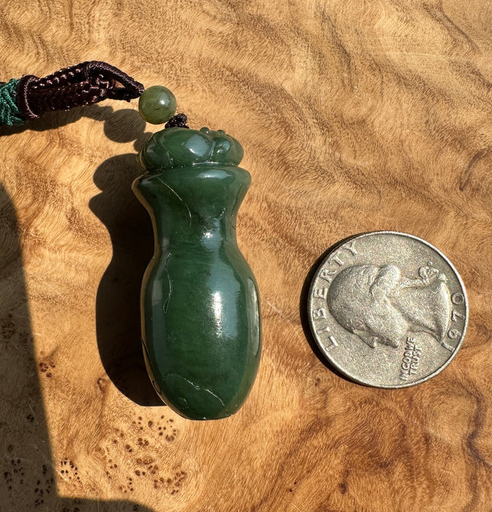 A polished dark green jade pendant shaped like a small bottle or vase, shown on a wood surface next to a U.S.