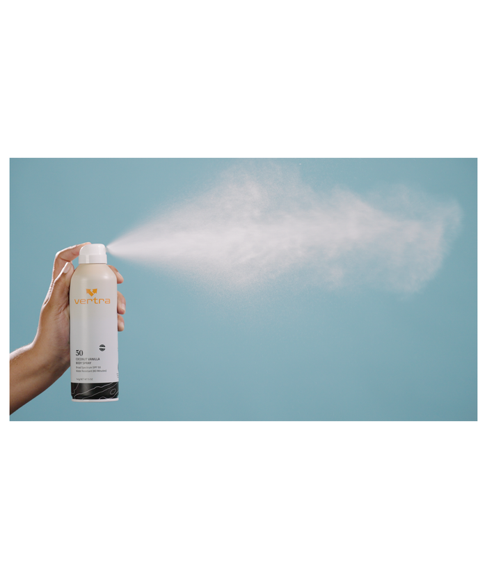 Hand holding and spraying Coconut Vanilla Sunscreen Body Spray SPF 50 | Vertra with fine mist visible against a blue background, showcasing skincare product application.