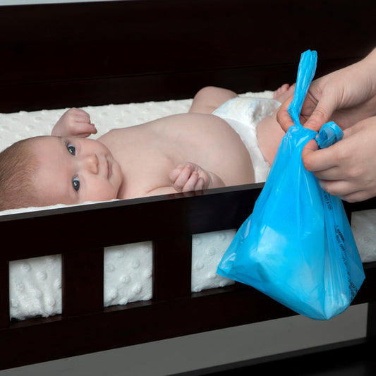 A baby in a diaper lies on a changing table as an adult uses Nuby Diaper Sacks (50 count), likely containing a used diaper for disposal.