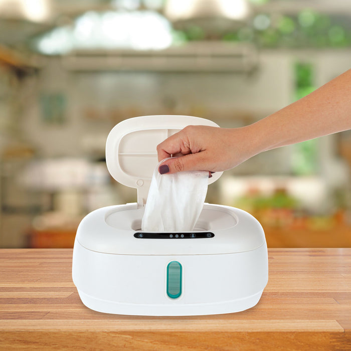 A hand pulls a wet wipe from the Nuby Hands-Free Wipe Warmer, featuring a green button and sitting on a wooden surface in a bright, blurred kitchen.