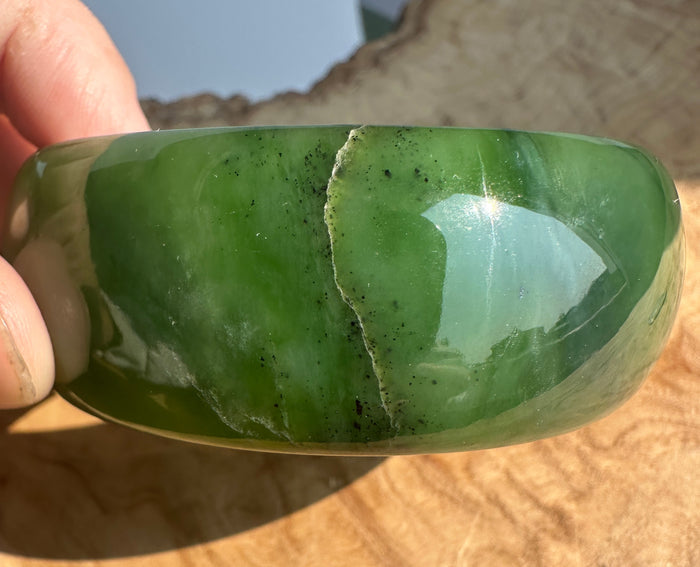 A hand holding a polished green jade bangle bracelet against a natural wood and outdoor background.