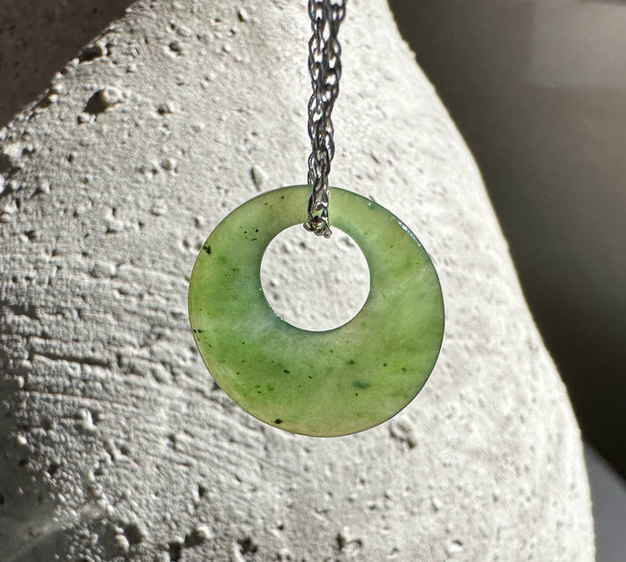 Pale green jade donut pendant hanging on a dark chain against a light textured stone surface