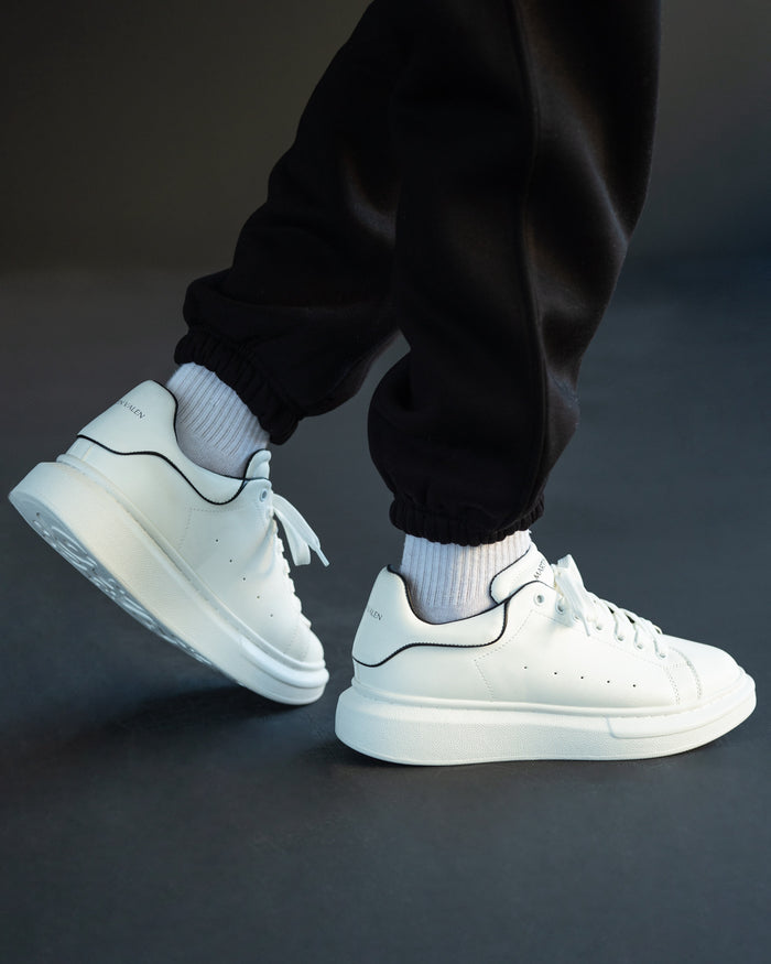 Martin Valen men's chunky white sneakers worn in a side walking pose with black joggers, showing black piping and logo on heel