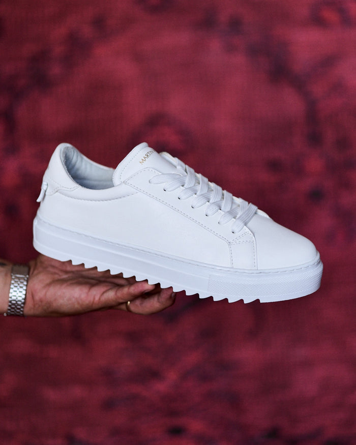 Martin Valen men's low-top white sneakers, close angled view of upper and laces with gold logo on the tongue against a red backdrop.