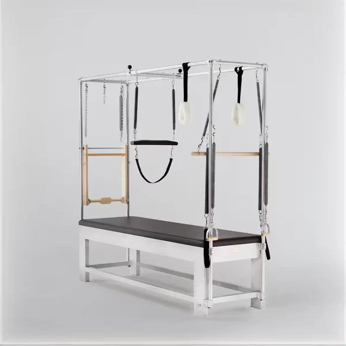 Pilates Cadillac reformer with silver aluminum frame, dark upholstered mat, trapeze bar, push-through bar, and adjustable spring attachments.