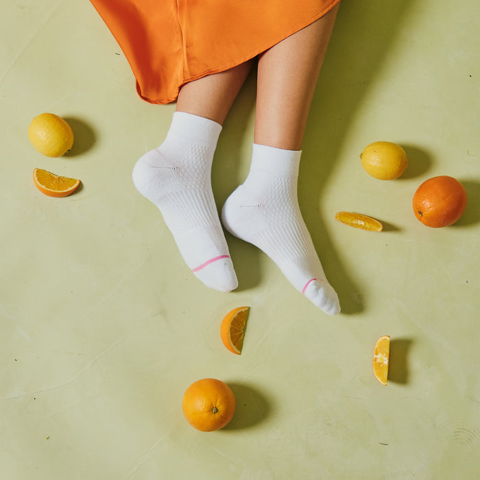 White quarter compression socks with pink accents, worn on feet surrounded by citrus fruits on a light green surface.