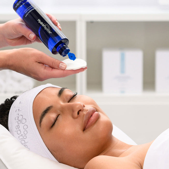 A person with a HydroPeptide headband lies with eyes closed while a hand pours Pre-Treatment Toner | HydroPeptide onto a cotton pad for skincare preparation.