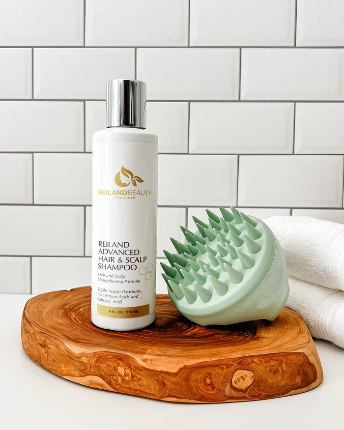 Reiland Advanced Hair & Scalp Strengthening Shampoo – 6 oz bottle displayed with soft green scalp massager on a natural wood spa tray. Formulated with Triple Action Postbiotic, Oat Amino Acids, and Salicylic Acid for clean, healthy scalp and thicker-looking hair.