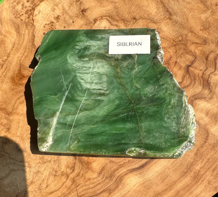 A raw green Siberian jade stone slab with natural marbling and white crystalline edges, labeled 'SIBERIAN' on a white sticker, displayed on a textured wood background.