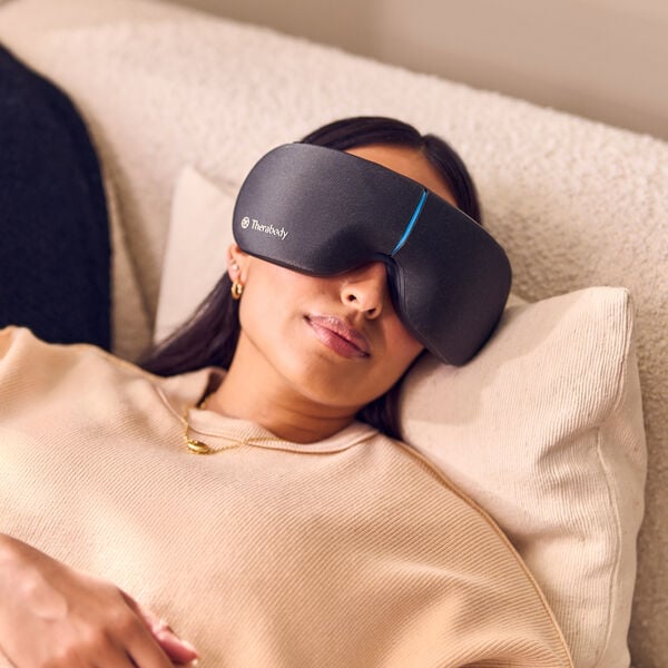 A woman relaxing on a couch wearing SmartGoggles 2 | Therabody, a wellness device designed for eye recovery and relaxation.