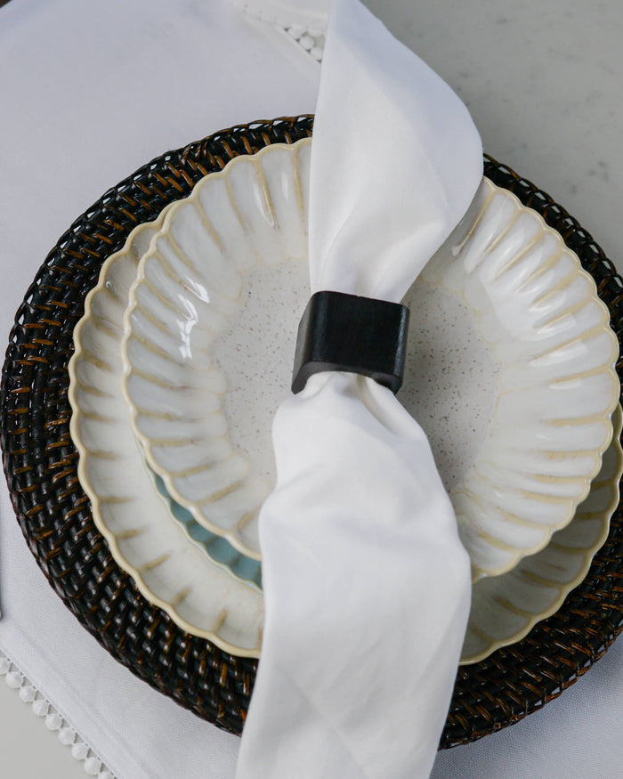 White ceramic scalloped pasta dish with a dark wood napkin ring on a white cotton table runner