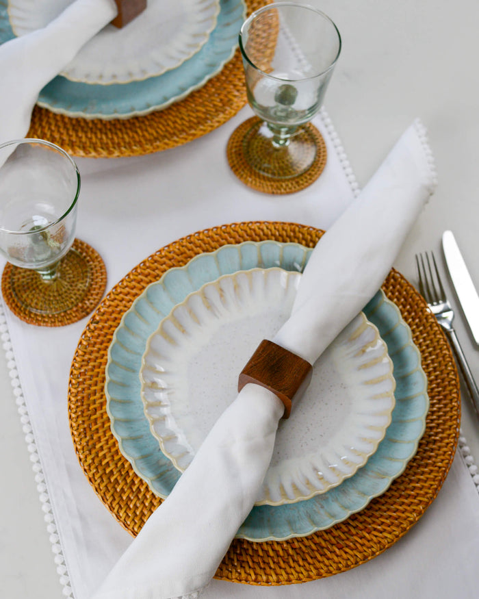 Dining table setting with ceramic plates, wooden napkin rings, woven rattan placemats, and cutlery on a white table runner.