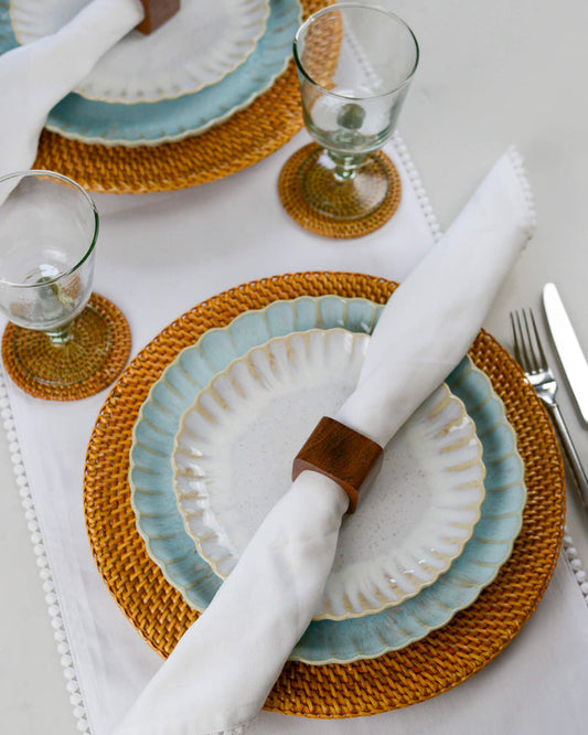 Dining table setting with ceramic plates, wooden napkin rings, woven rattan placemats, and cutlery on a white table runner.