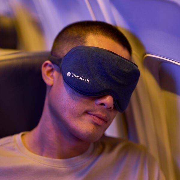 Man wearing a black Therabody Sleepmask | Therabody, resting comfortably on an airplane seat, promoting relaxation and wellness during travel.
