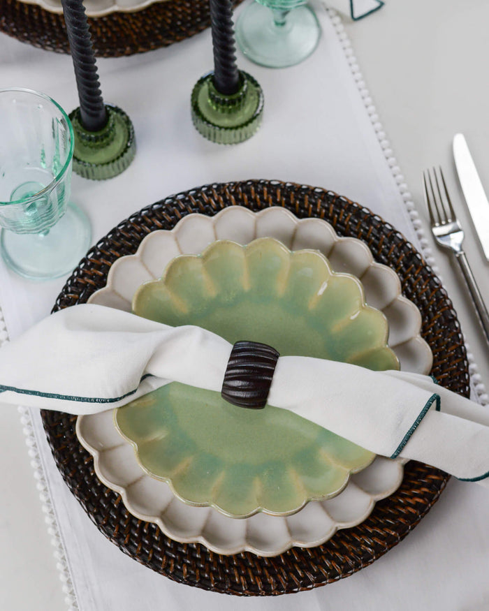 Decorative place setting with a scalloped-edge plate, napkin, dark wooden napkin ring, and glasses on a white tablecloth.
