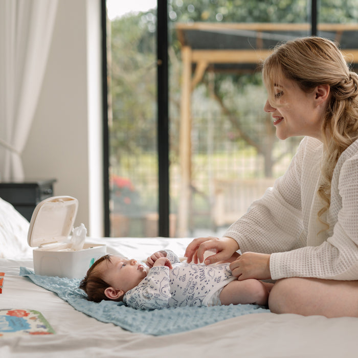 A smiling woman changes a baby’s diaper on a blue mat, with the Nuby Wipe Dispenser nearby. The dispenser features a weighted plate. Books rest close by as sunlight pours through large windows in the background.