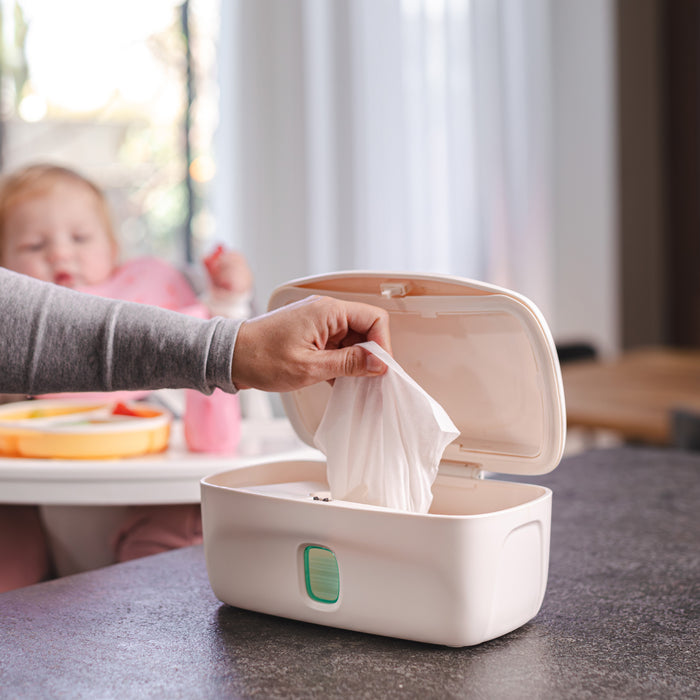 A hand pulls a wet wipe from the Nuby Wipe Dispenser with a weighted plate on the kitchen counter, while a baby sits in a high chair—ideal for quick diaper changes.